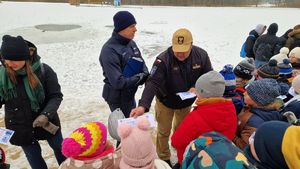 Policjant omawia tematykę bezpieczeństwa, strażak rozdaje uczestnikom ulotki.