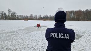 Policjant stoi przed zamarzniętym zbiornikiem wodnym i przygląda się strażakowi znajdującemu się w przeręblu.