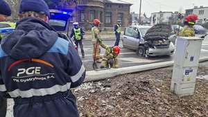 Miejsce zdarzenia drogowego, na którym widać uszkodzoną latarnię, rozbite auto i służby pracujące.