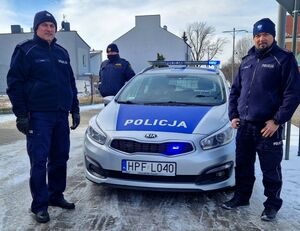 Dzielnicowi stoją obok radiowozu, z tyłu znajduje się strażnik miejski.