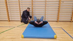 Mężczyzna wykonuje siady z leżenia na plecach. Czynność nadzoruje umundurowany policjant.