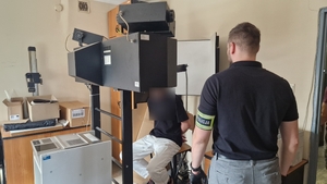 Nieumundurowany policjant stoi obok podejrzanego mężczyzny. Podejrzany siedzi na urządzeniu do fotografii.
