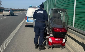Policjantka stoi prze mężczyźnie siedzącym w czerwonym, elektrycznym wózku inwalidzkim. Oboje znajdują się na drodze ekspresowej, w miejscu gdzie prowadzona jest interwencja.