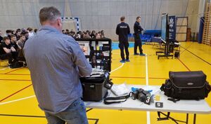 Technik kryminalistyki stoi przy stoisku ze sprzętem wykorzystywanym w codziennej służbie. W tle widać uczniów zgromadzonych na spotkaniu.