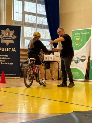 Zawodnik pokonuje rowerem tor przeszkód. Mężczyzna stoi obok zawodniczki i wskazuje jej palcem element toru.
