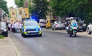Policjanci w radiowozie i funkcjonariusz na motocyklu zabezpieczający przejście procesji.