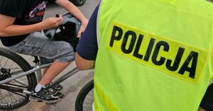 Chłopiec siedzi na rowerze i trzyma kask w ręku. Obok niego stoi policjant w odblaskowej kamizelce.