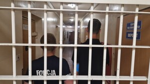 policjant z osobą zatrzymaną stoją tyłem za kratami.
