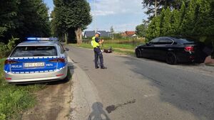 umundurowany policjant wykonuje oględziny zaparkowanego pojazdu marki BMW. w tle za umundurowanym policjantem leży wywrócony motocykl. widać również policyjny radiowóz