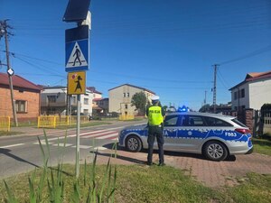 umundurowany policjant stoi przy oznakowanym radiowozie w rejonie szkoły i kontroluje nieprawidłowości w oznakowaniu.