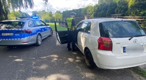 umundurowany policjant stoi przy kontrolowanym pojeździe. w tle stoi w poprzek drogi radiowóz oznakowany na sygnale