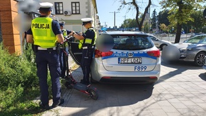 umundurowani policjanci z wydziału ruchu drogowego w kamizelka odblaskowych kontrolują kierującego 14-latka hulajnogą elektryczną.