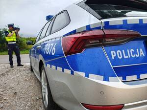 radiowóz policyjny, a w tle umundurowany policjant dokonuje pomiaru prędkości