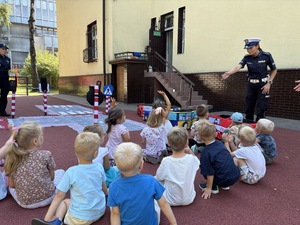 Policjantki rozmawiają z grupą przedszkolaków o bezpieczeństwie na drodze, w tle jest też mata edukacyjna autochodzik