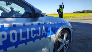 oznakowany radiowóz policji a w tle policjantka w kamizelce odblaskowej wydaje polecenia do zatrzymania się pojazdu w celu przeprowadzenia kontroli